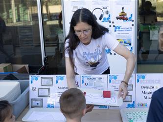 A woman with a white shirt and glasses leans over a table to help children.
