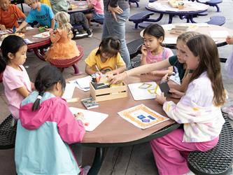A group of children gather around a table outside to work on a collective art project. 