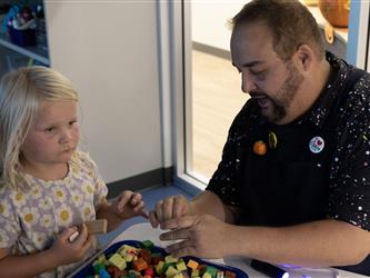 A man in a black shirt shares colorful blocks with a girl with blonde hair.