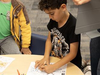 A boy writes on a piece of paper standing next to a table. 