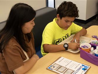 A boy in a yellow shirt is drawing while his mother looks on. 